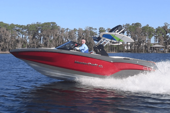 Red motorboat speeding on a lake with a wakeboarder in tow, trees in the background.