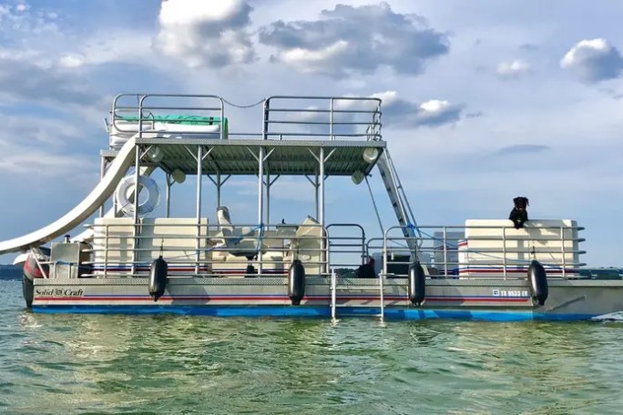 Pontoon boat with a slide on a lake under a cloudy sky.