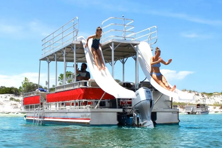 Two women sliding down slides on a pontoon boat in clear water.
